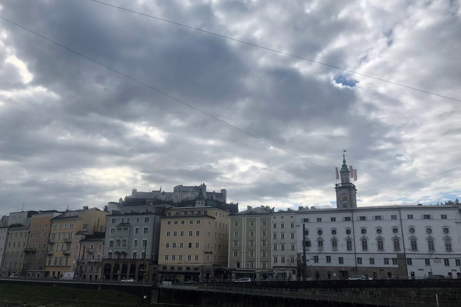 Zu sehen ist eine Häuserfront in Salzburg. Im Hintergrund ist ein Kirchturm und eine Burg zu sehen. Der Himmel ist bewölkt, aber darüber leicht bläulich.