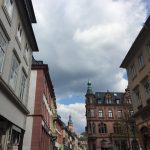 Die Heidelberger Innenstadt gesehen von der Hauptstraße. Auf dem Bild sind neben vielen Steinhäusern auf die katholische Kirche, viele Wolken und kleine Teile des Heidelberger Schloßes zu sehen.